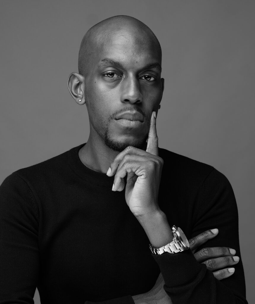 Black-and-white headshot of Jamell Henderson, a bald man wearing a watch and resting his chin on his hand with a thoughtful expression, emphasizing leadership and reflective charisma.