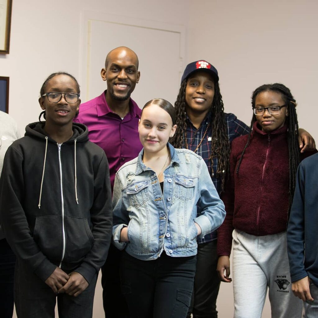 Jamell Henderson in a purple shirt stands among a diverse group of teens, all smiling in casual attire, highlighting youth community engagement.