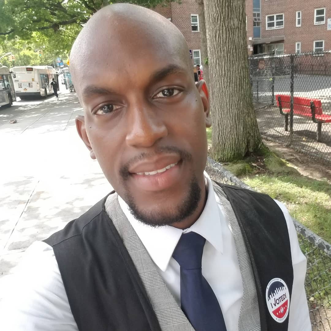 A smiling Jamell Henderson in a black vest and tie with an “I Voted” sticker, takes a selfie on a sunny NYC sidewalk near a bus stop and brick apartment building.