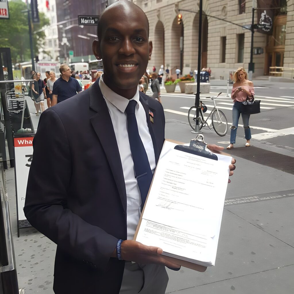 Jamell Henderson in a navy suit smiling on a busy New York City street, holding a clipboard with campaign paperwork for a City Council run, with pedestrians and urban architecture in the background.